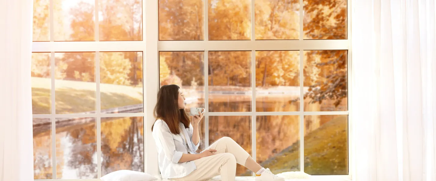 Offre_fentre_tryba.webp Une femme assise sur un banc devant une grande fenêtre, entourée d'un paysage d'automne, savourant une tasse de café.