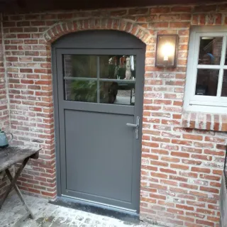 porte_belgique.webp Une porte grise moderne avec une fenêtre en verre est installée sur un mur en briques rouges, éclairée par une appliqué lumineuse.