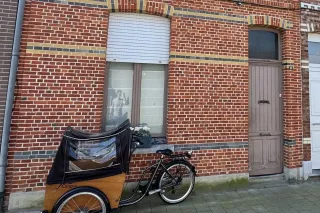 porte_entree_fenetre_belgique.webp Un vélo cargo est stationné devant une maison en brique rouge avec un volet mi-fermé et une porte en bois.