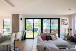 TRYBA_baie_vitre_coulissante_gris-anthracite-avec-brise-soleil-orientable.webp Un salon moderne et lumineux avec un canapé beige, des coussins colorés et de grandes baies vitrées donnant sur un jardin.