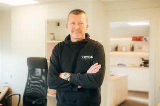 tryba_belgique_contactez_nous.webp Un homme en sweat-shirt 'TRYBA' se tient avec les bras croisés dans un bureau lumineux et moderne.