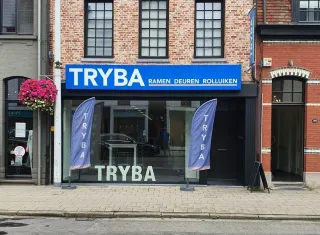 TRYBA_Belgique,_une_histoire_alliant_passion_et_expertise.webp Façade d'un magasin Tryba avec une enseigne bleue et des drapeaux publicitaires, spécialisée dans la vente de fenêtres, portes