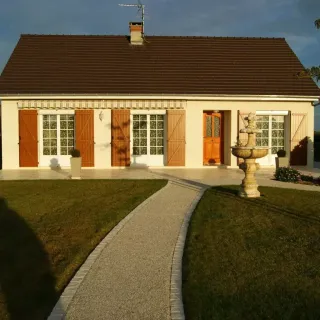 TRYBA_volet_battant.webp Une charmante maison de plain-pied avec volets en bois, allée en gravier et fontaine dans le jardin.