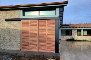 tryba_volet_sublime.webp Façade moderne avec volets en bois clair et murs en pierre naturelle, intégrée dans un environnement résidentiel.