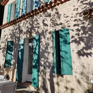 volet_battant_orientable.webp Façade ensoleillée d'une maison avec des volets bleus et des ombres d'arbres projetées sur le mur.