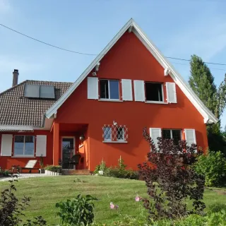 volet_battant_pvc.webp Maison rouge avec volets blancs dans un jardin, sous un ciel bleu.