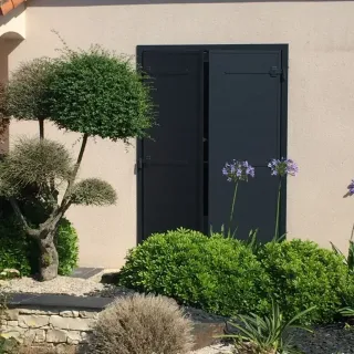 volets_battants_motorisation.webp Façade de maison avec volets noirs fermés, entourée de plantes et d'un jardin bien entretenu.