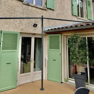volets_battants_personnalisation.webp Façade de maison avec volets verts et terrasse en bois, ornée d'une grande plante en pot.