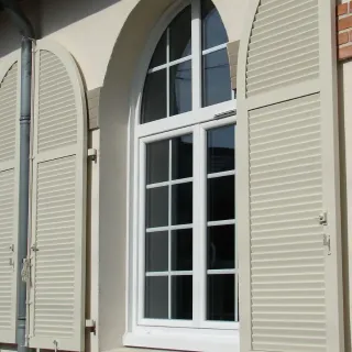 volets_battants_securisee.webp Fenêtre en PVC blanc à double vitrage avec volets en bois clair, offrant une touche élégante et fonctionnelle à la façade.
