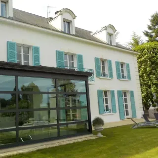 volets_battants_securite.webp Grande maison blanche avec volets bleus et véranda moderne donnant sur un jardin verdoyant.